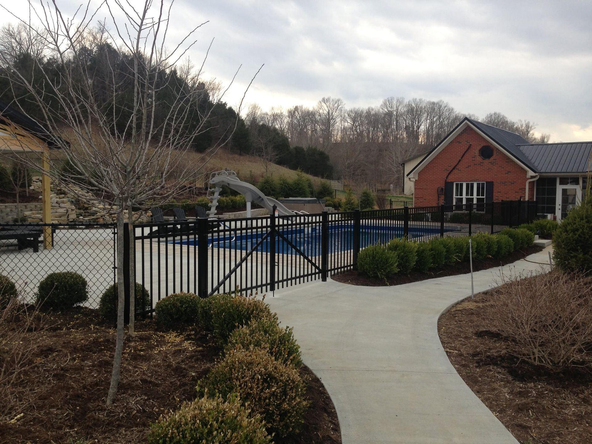 Curved walkway beside an outdoor pool, with black fence, shrubs, and brick buildings in the background