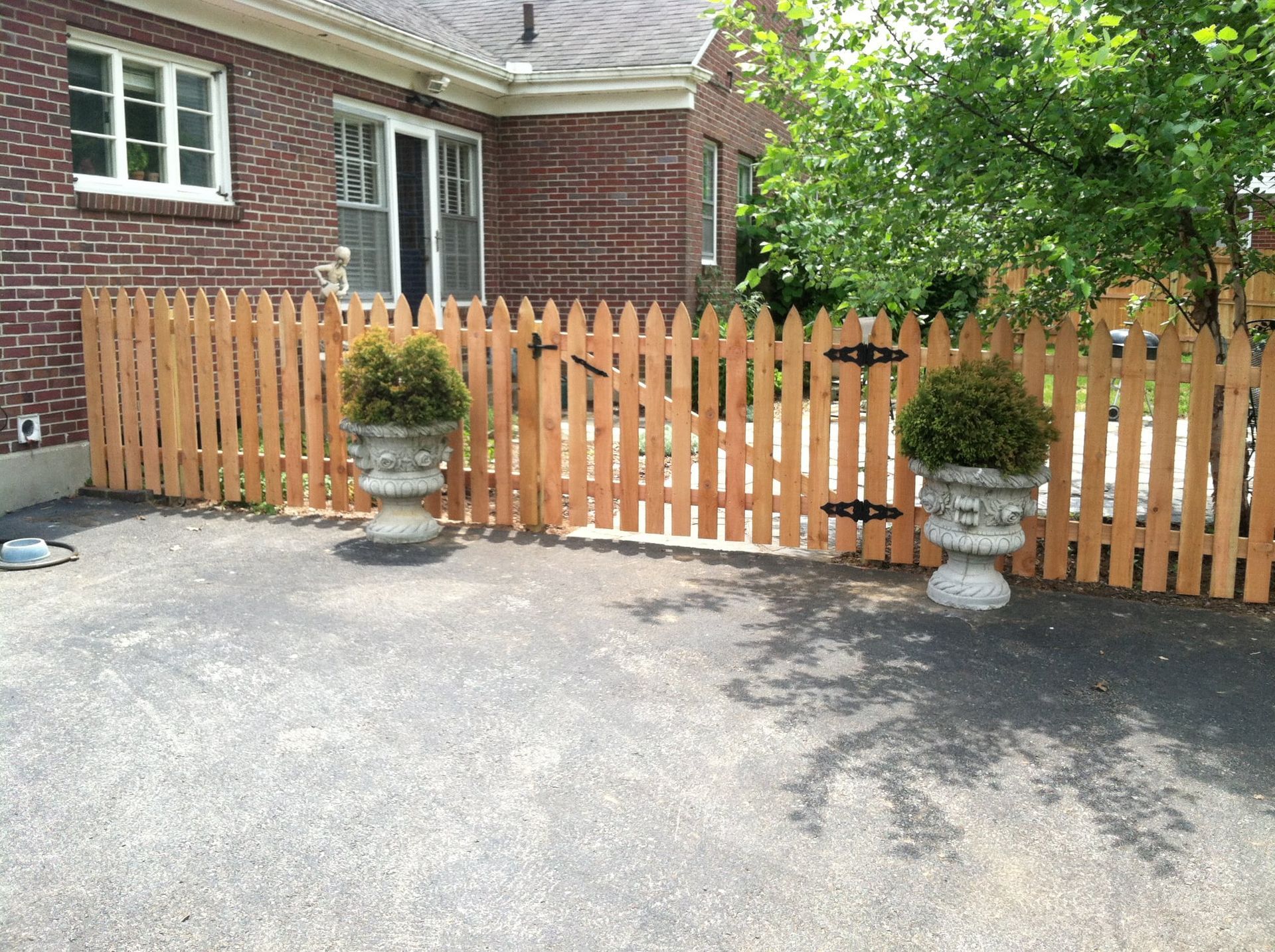 Brick house front yard with wooden picket fence, potted shrubs, and a paved driveway.