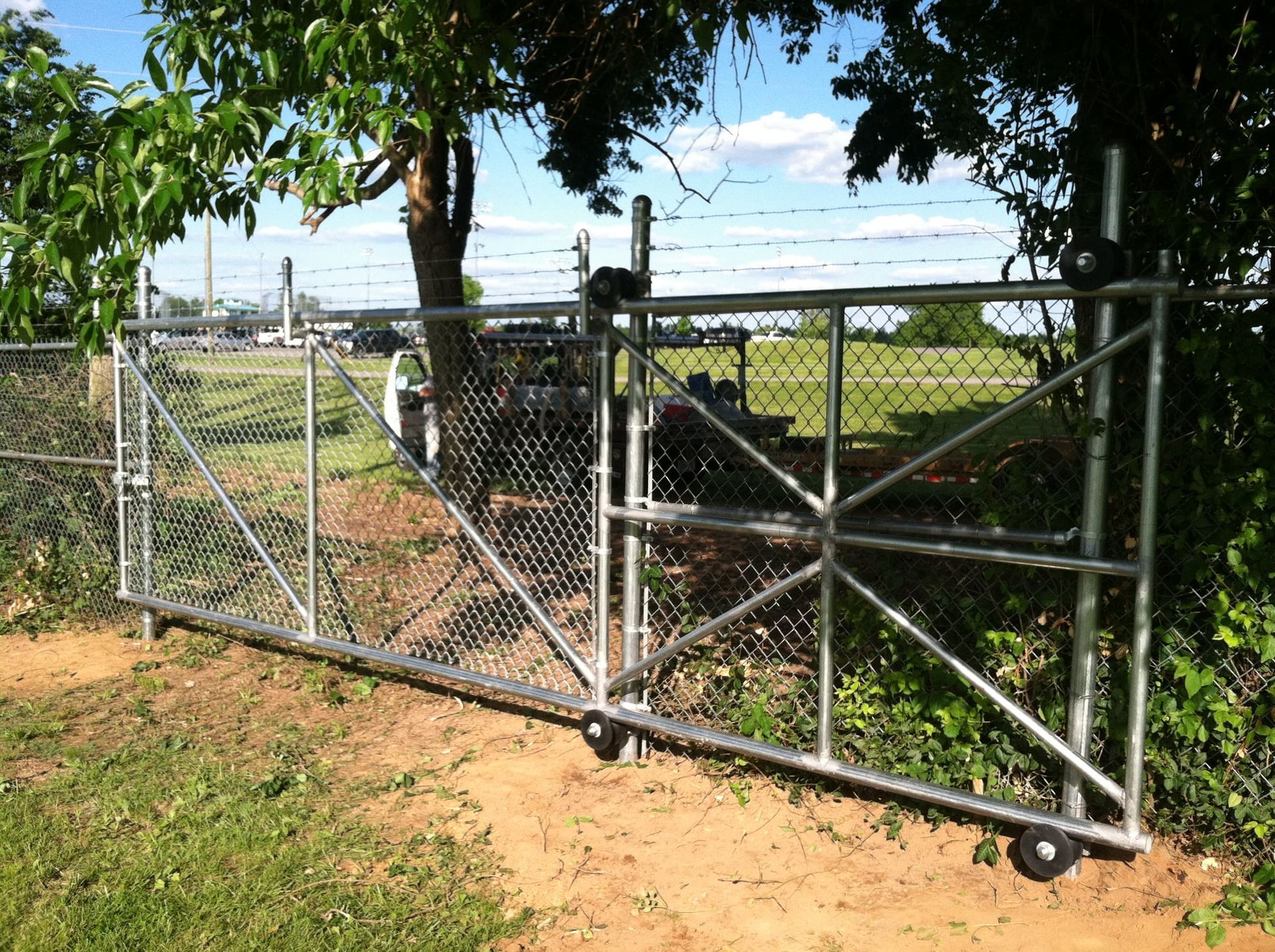 Chain-link farm gate with metal frame beside a dirt path and trees under a blue sky