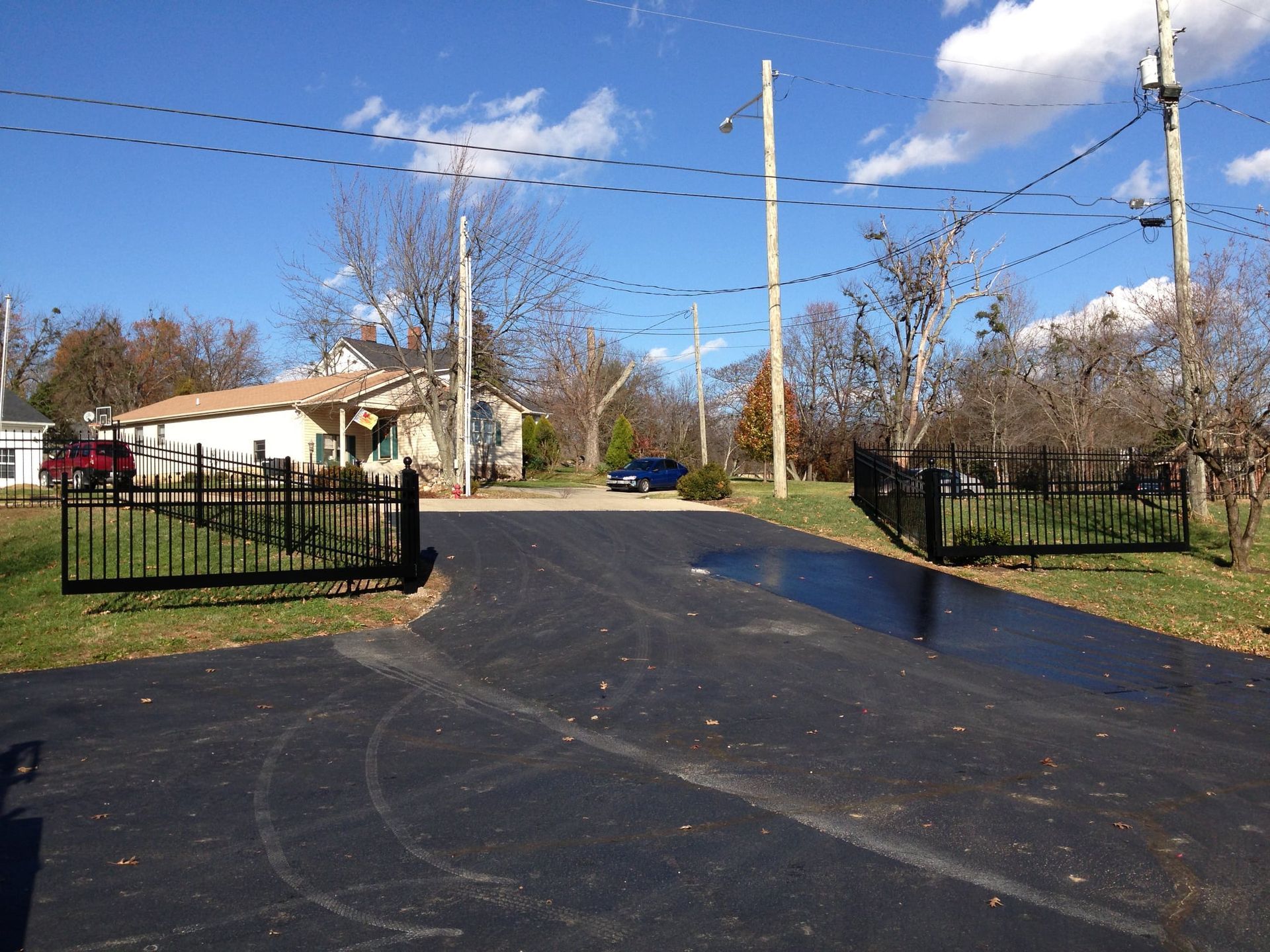 Driveway entrance with open black gates, utility poles, and a house under a clear blue sky
