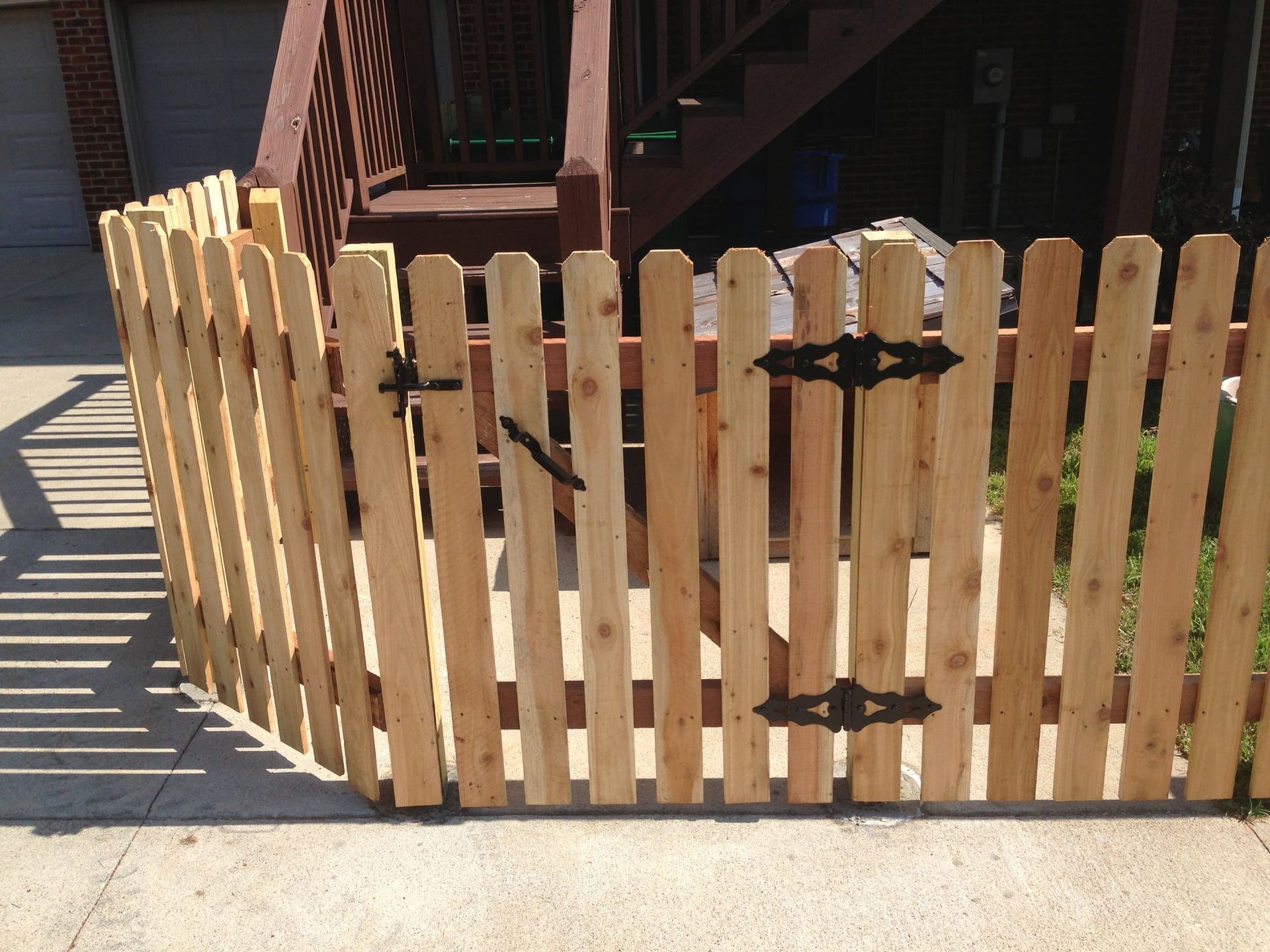 Wooden picket fence with black hinges and latch beside a porch and sidewalk in sunlight