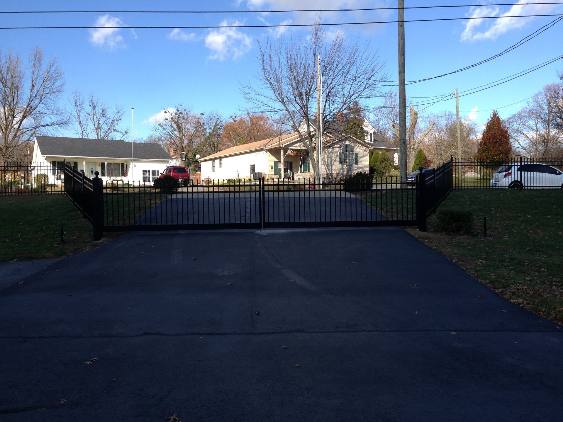 Black driveway leading to a closed metal gate, with houses, trees, and a blue sky beyond.