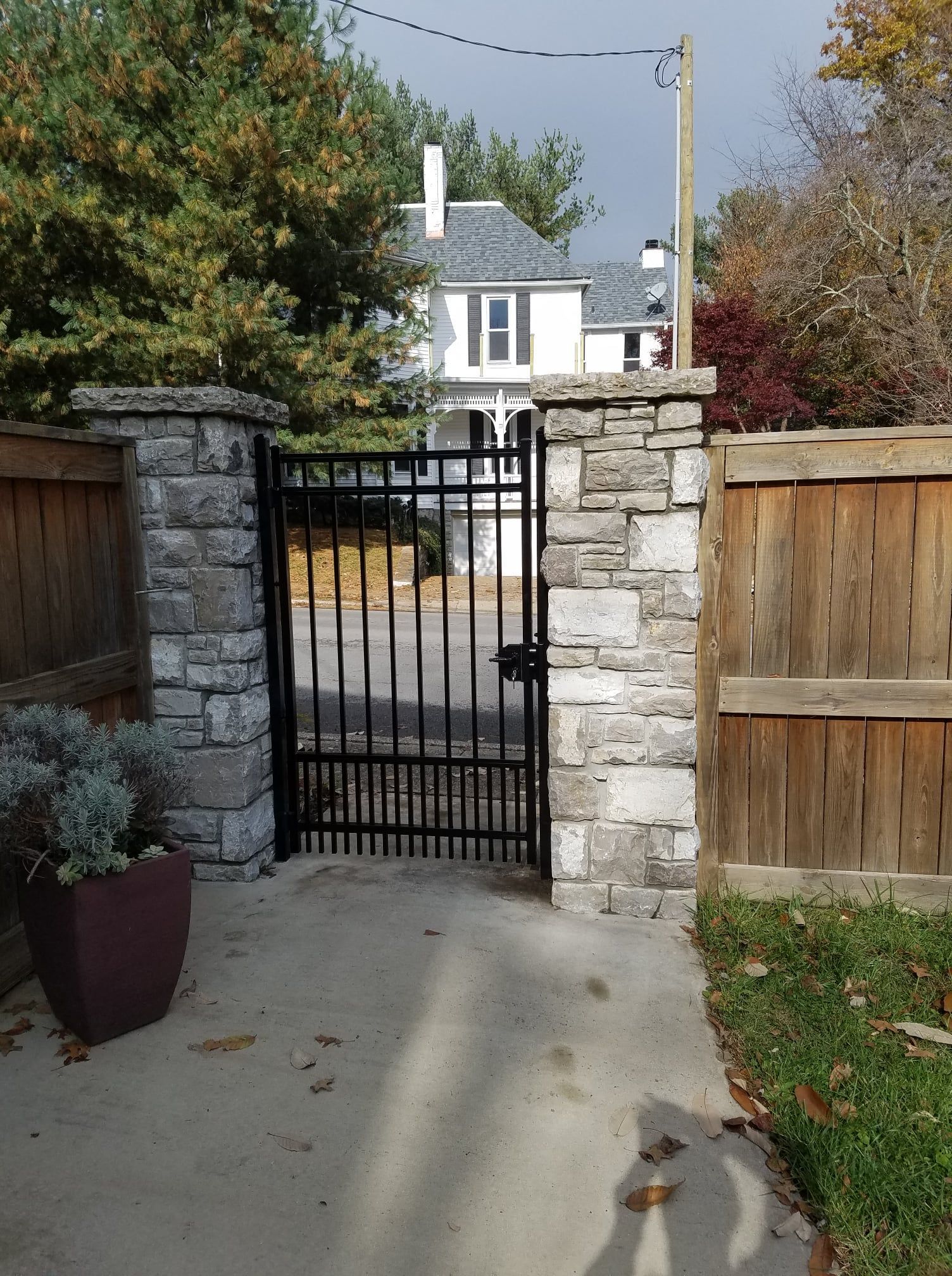 Front gate between stone pillars and wooden fence leading to a residential driveway, with a house beyond.