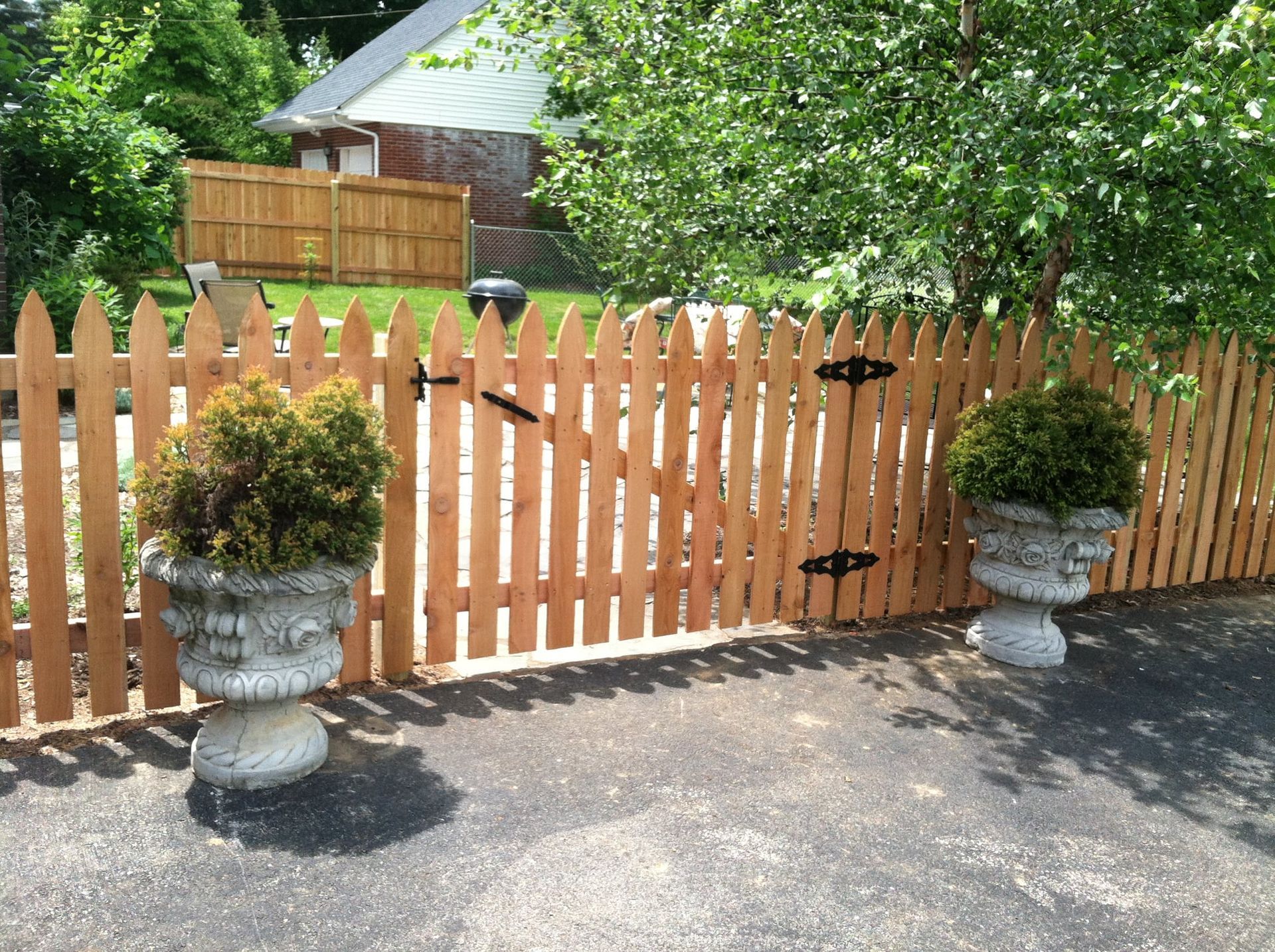 Wooden picket gate with black hinges between stone planters beside a driveway and leafy yard