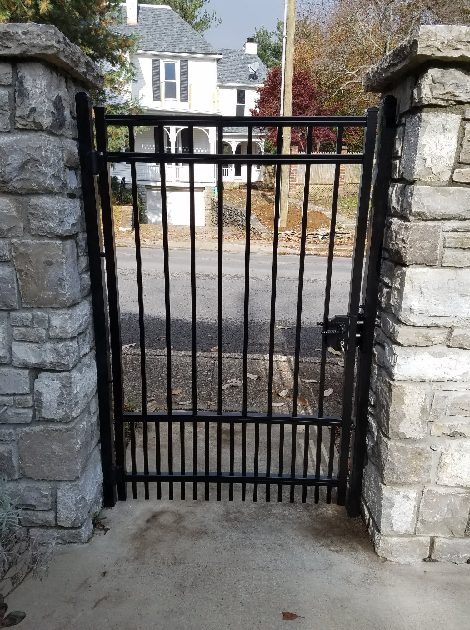 Black metal gate between stone pillars, opening onto a driveway with a house in the background.