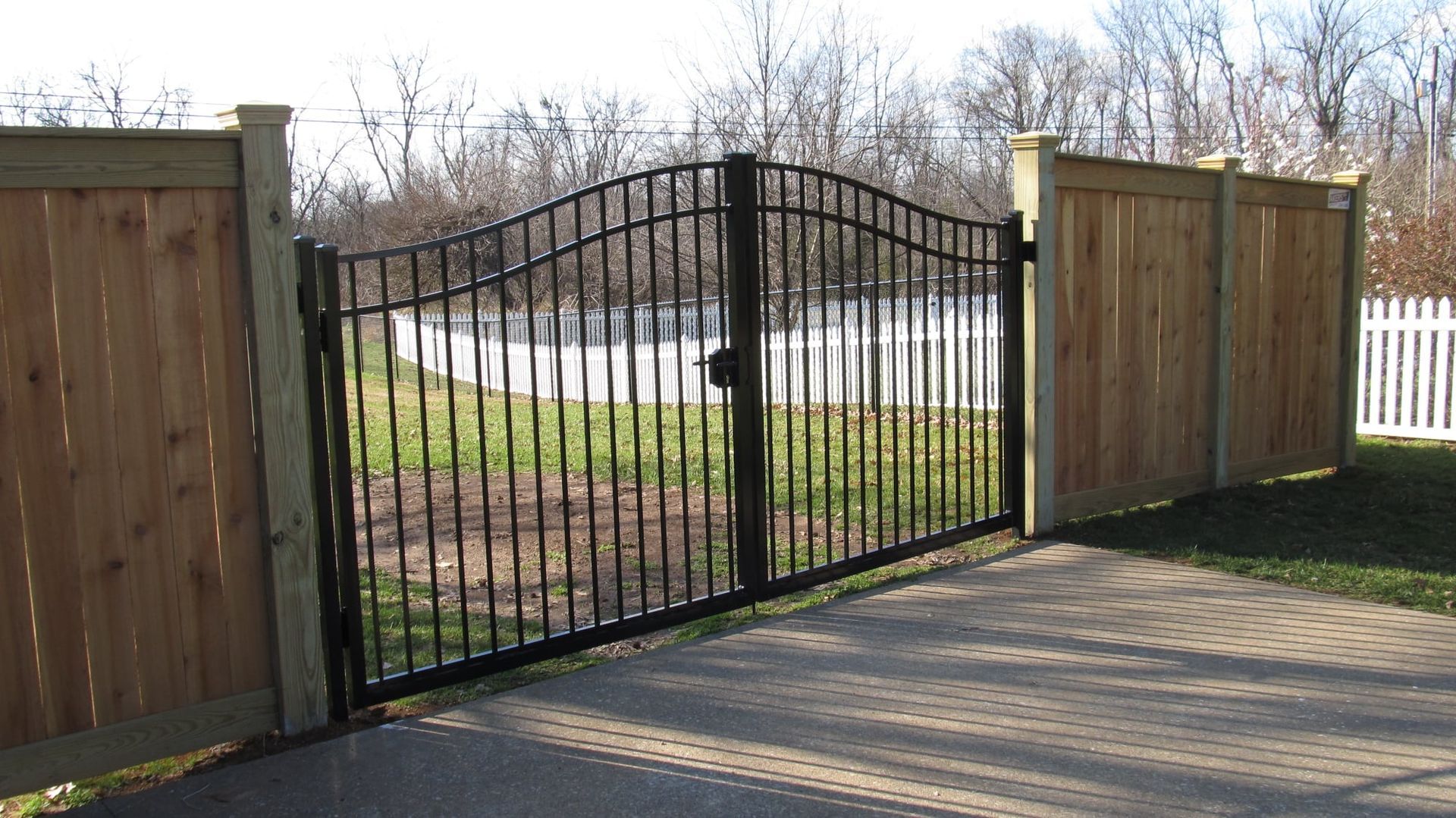 Black metal gate between wooden fences beside a paved driveway in a sunny yard