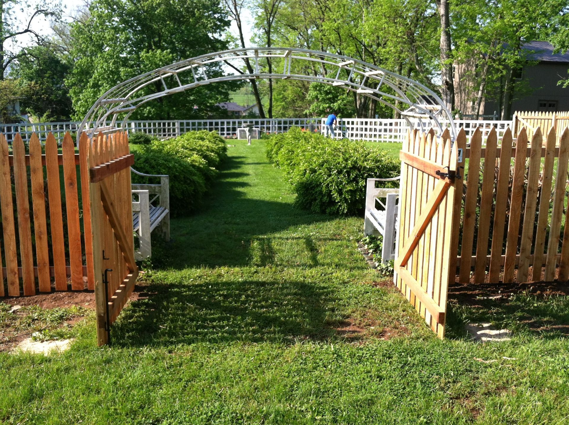 Open wooden gate leading to a grassy garden with white trellis archway and fences