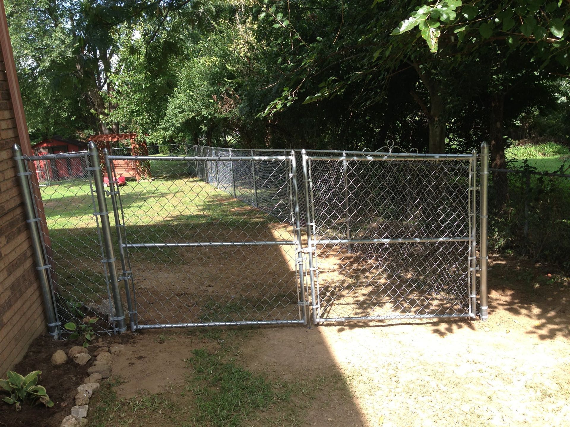 Chain-link gate and fence in a sunny backyard with trees and a brick wall.