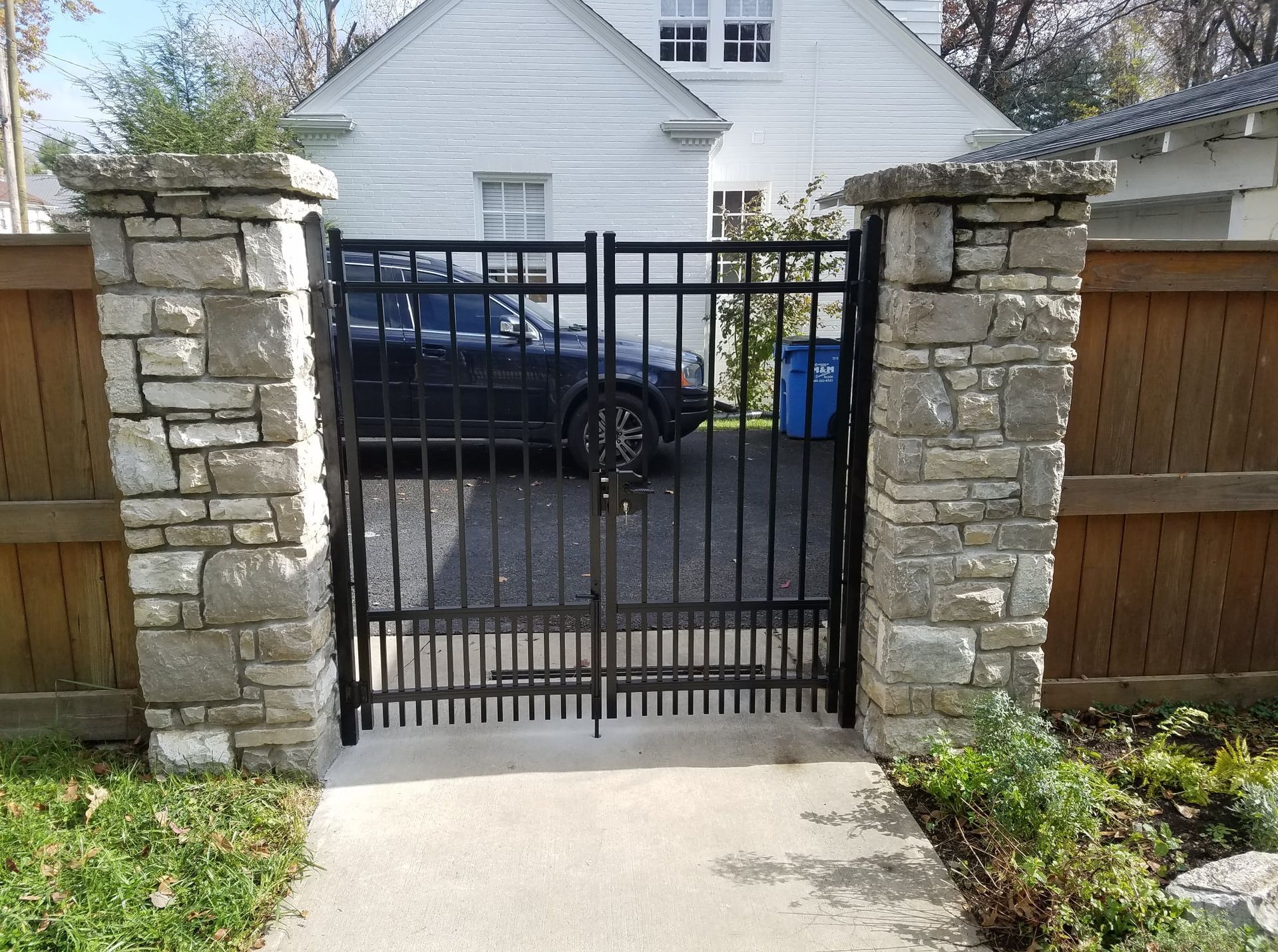 Black metal gate between stone pillars and wooden fence, opening to a driveway and white house.