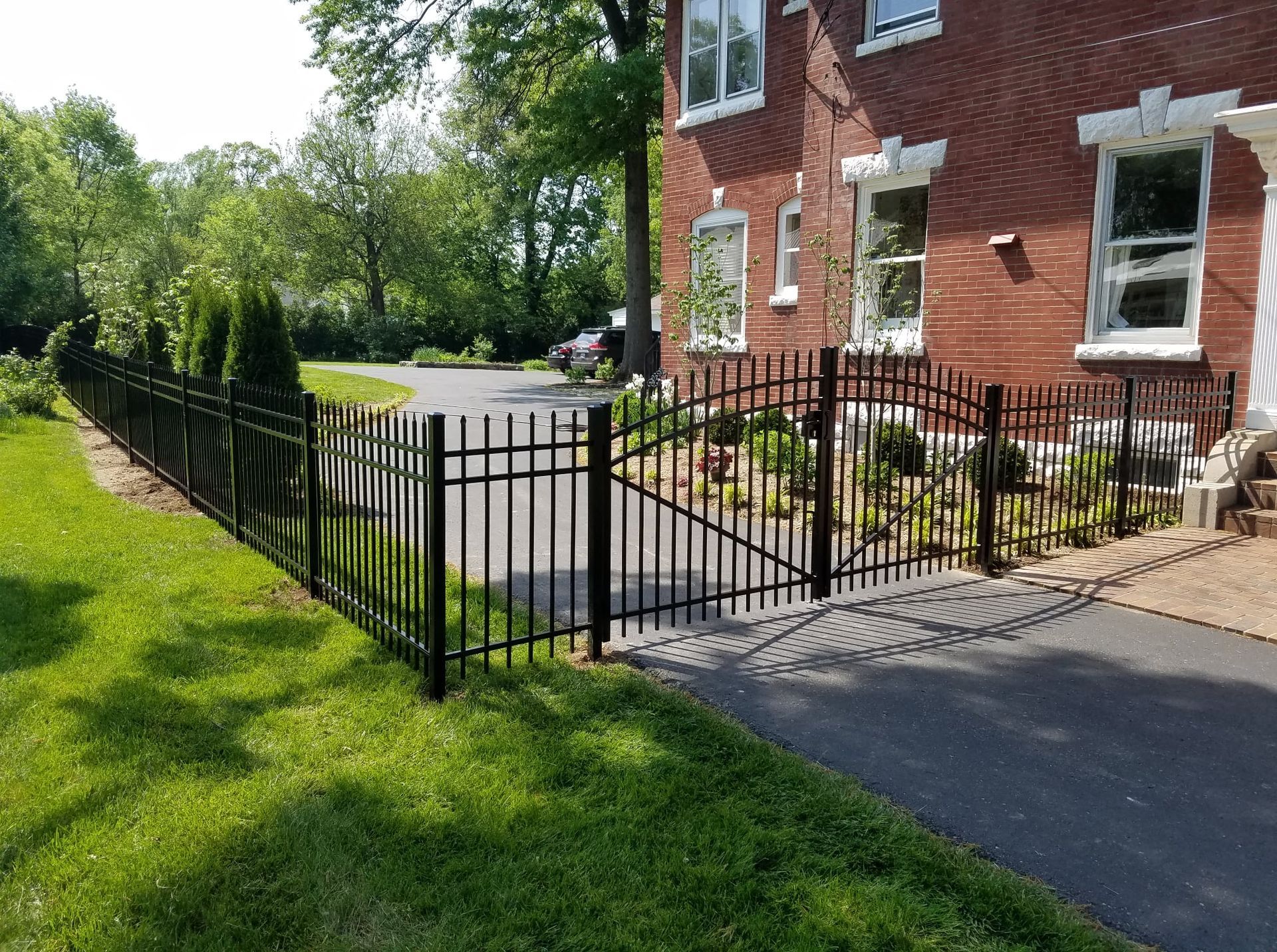 Black metal fence beside a brick house, with a garden and driveway in bright sunlight.