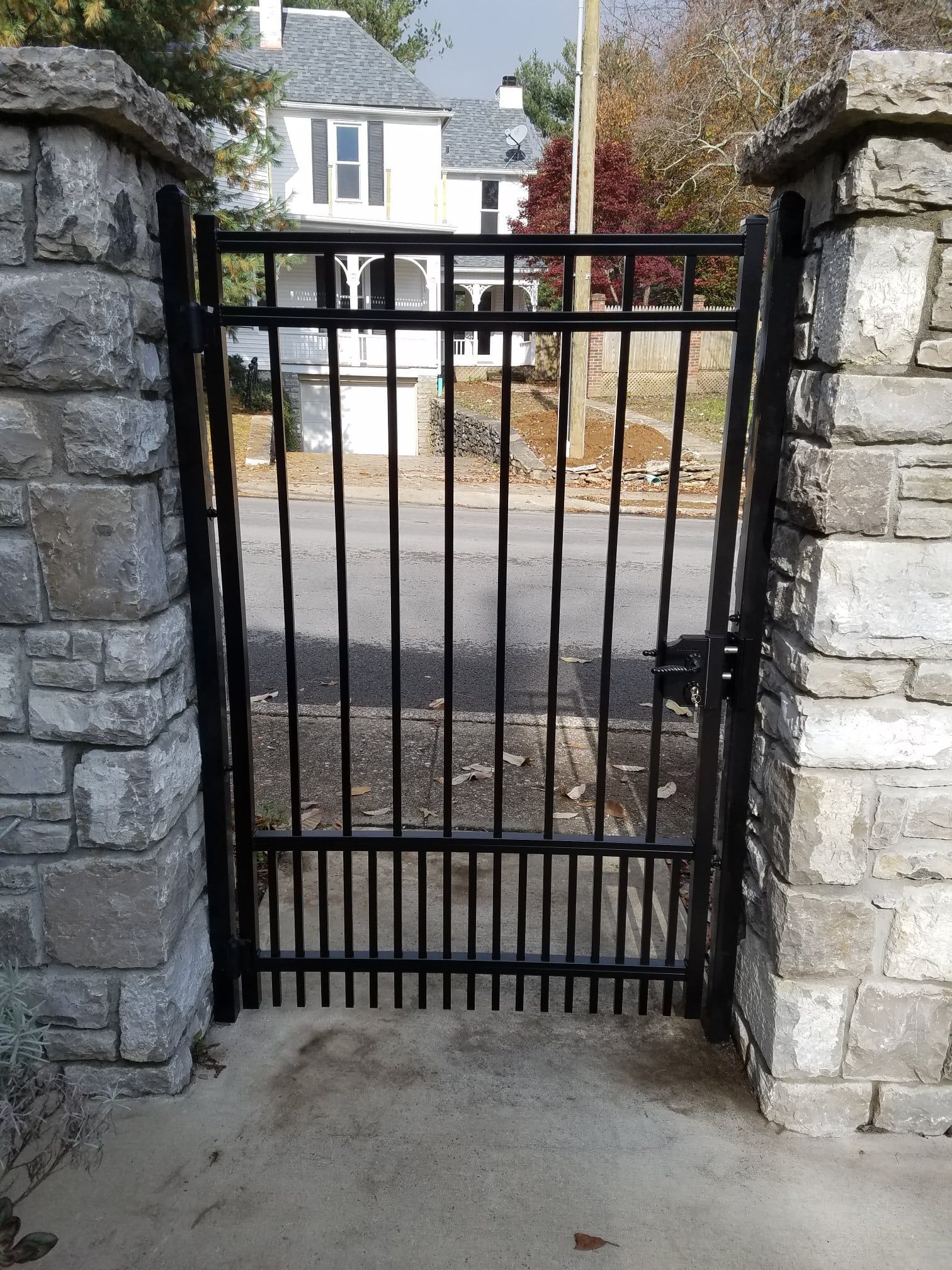 Black metal gate between stone pillars at a driveway entrance, with a house visible beyond.