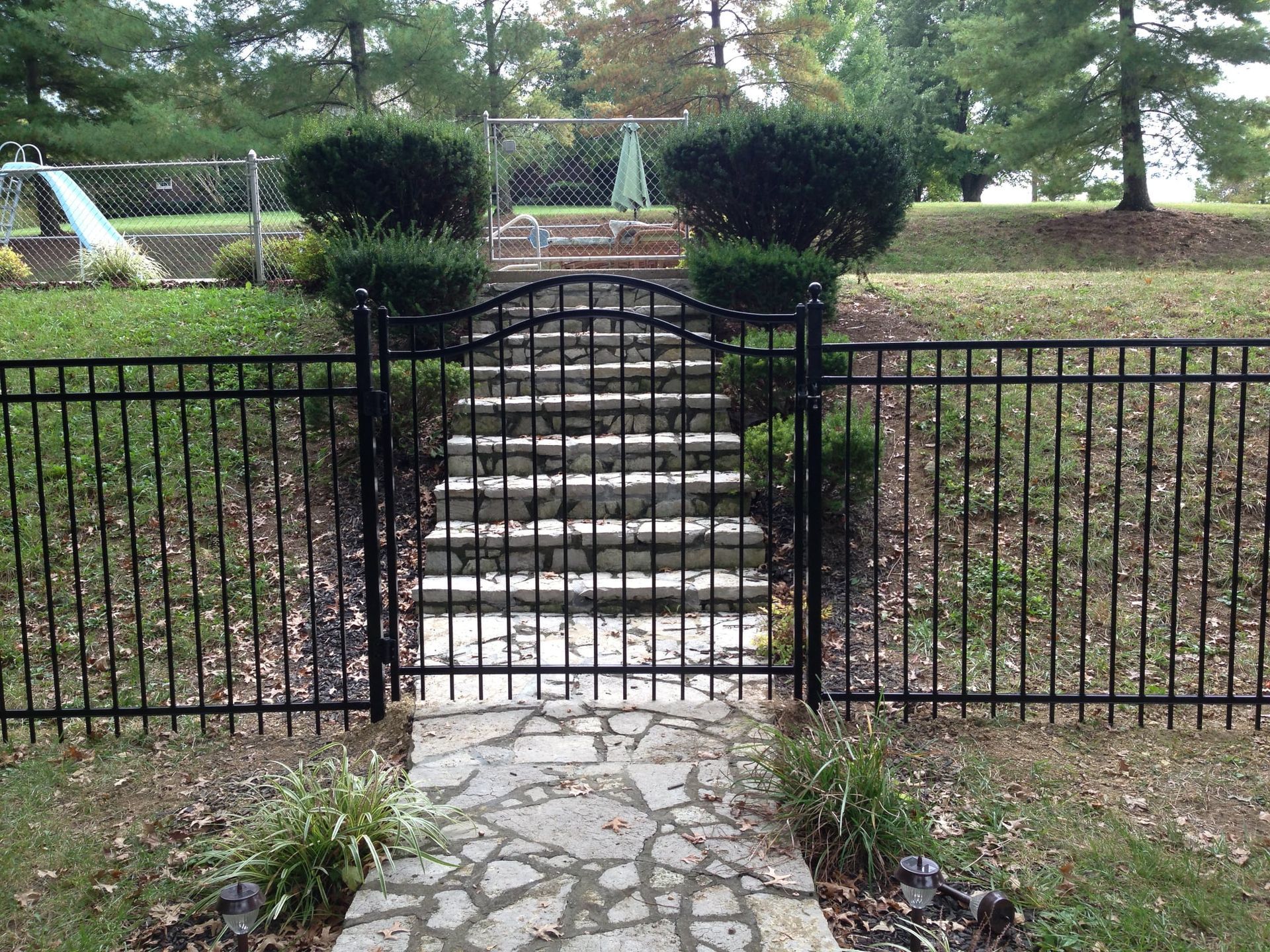 Black iron gate and stone steps leading to a park path, with trimmed bushes and trees behind.