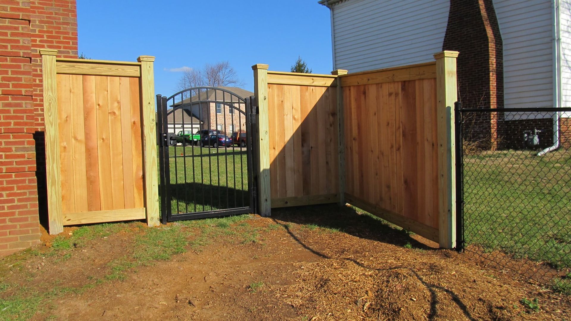 Open wooden gate leading to a black metal gate between brick houses over a dirt yard