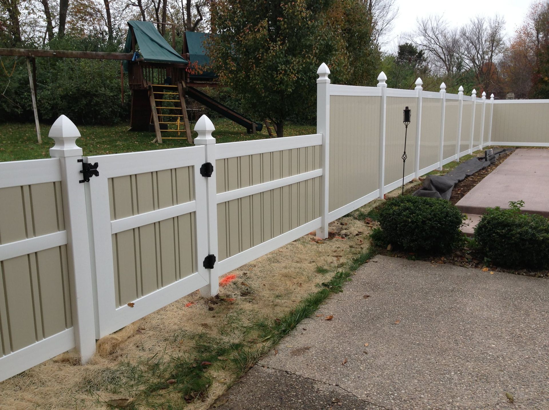 White vinyl fence along a gravel driveway and walkway in a yard with trees and a playset.