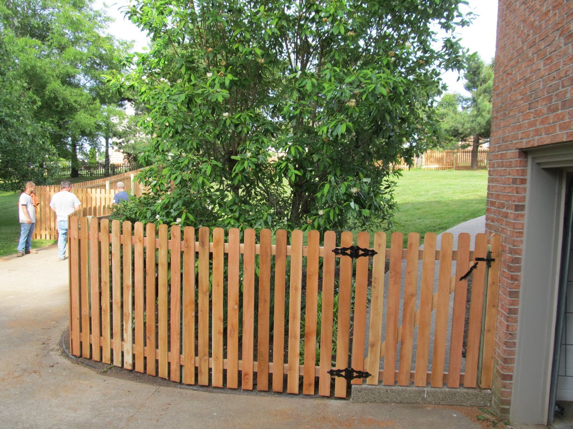Wooden gate in a driveway beside a brick house, with a green yard and trees behind it.