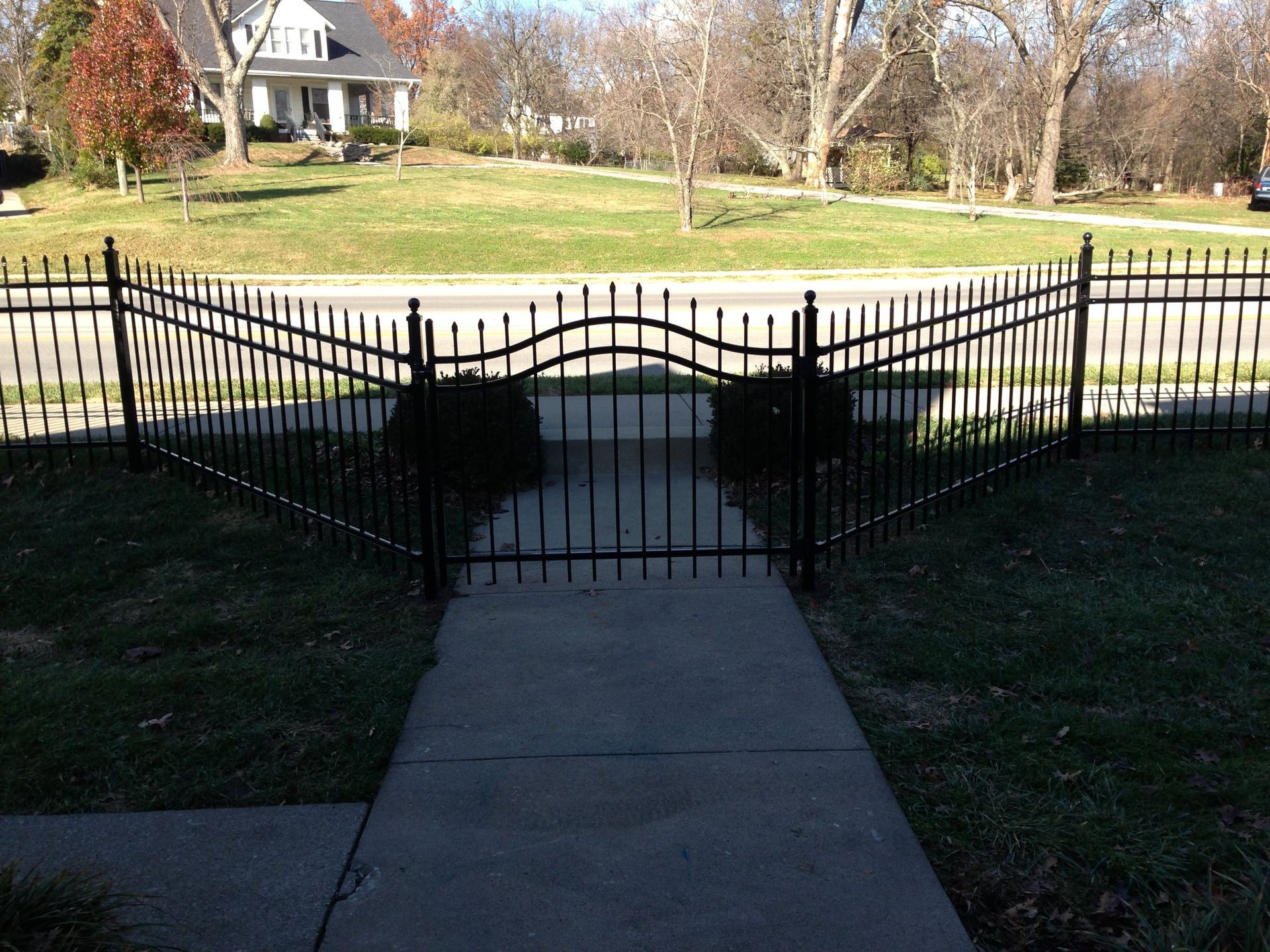 Black iron gate at the end of a concrete walkway, opening onto a grassy yard with a house in the background