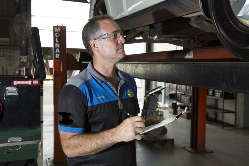 Mechanic in blue work shirt inspecting a vehicle on a lift with a clipboard in an auto shop — Auto Inspections & Road Services N.T. in Woolner, NT