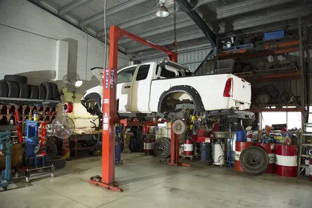 White pickup truck lifted on a hydraulic hoist in an auto repair shop with tires and tools nearby — Auto Inspections & Road Services N.T. in Woolner, NT