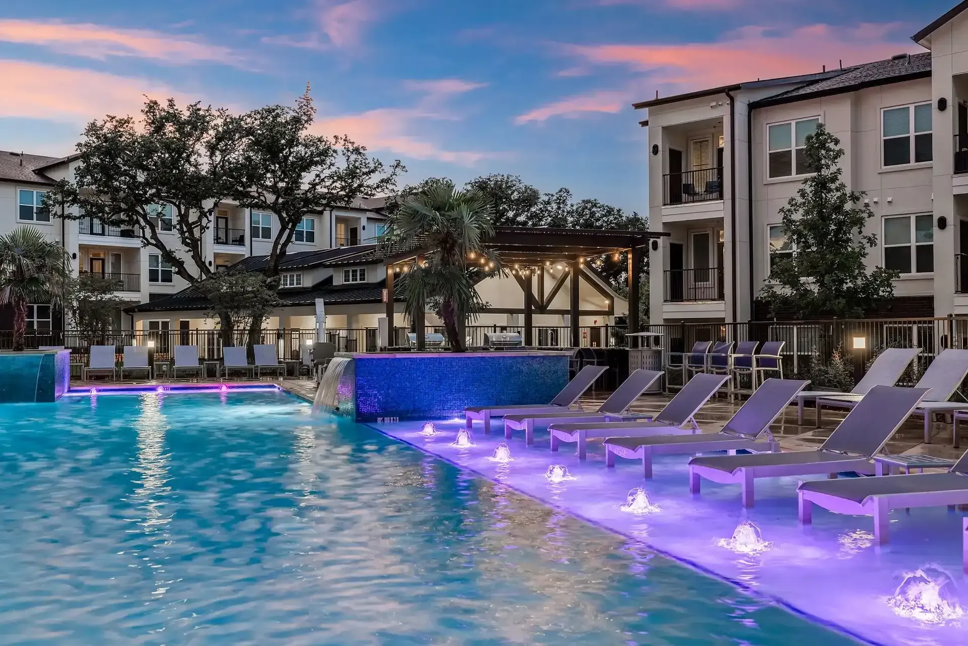 A large swimming pool with purple lights and lounge chairs in front of the apartment building at The Gabriel, which offers spacious apartments in Leander, TX, near Dell Technologies.