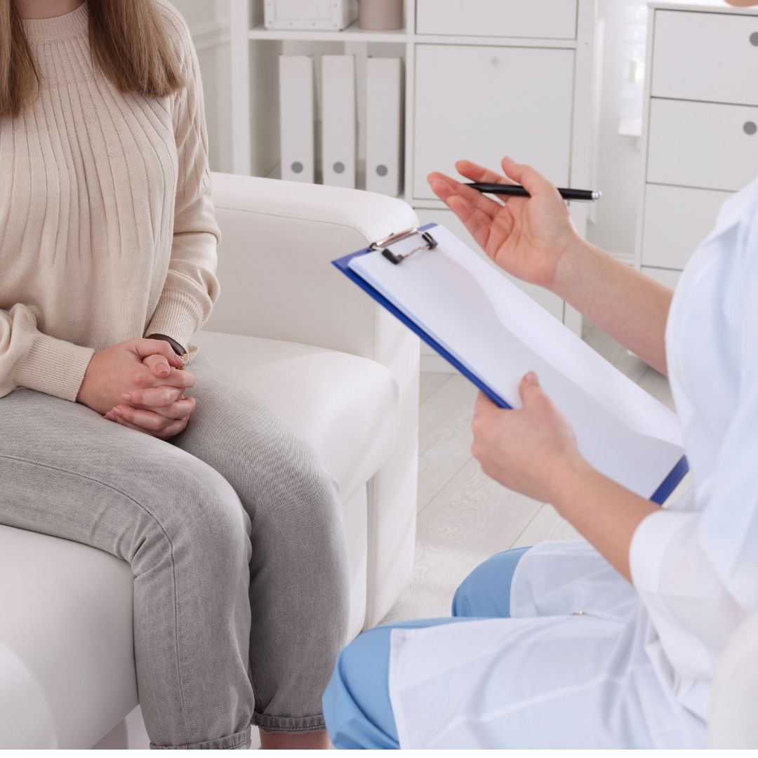 A woman in a light sweater sits on a white couch, talking with a medical professional holding a clipboard. The setting appears to be a doctor's office.