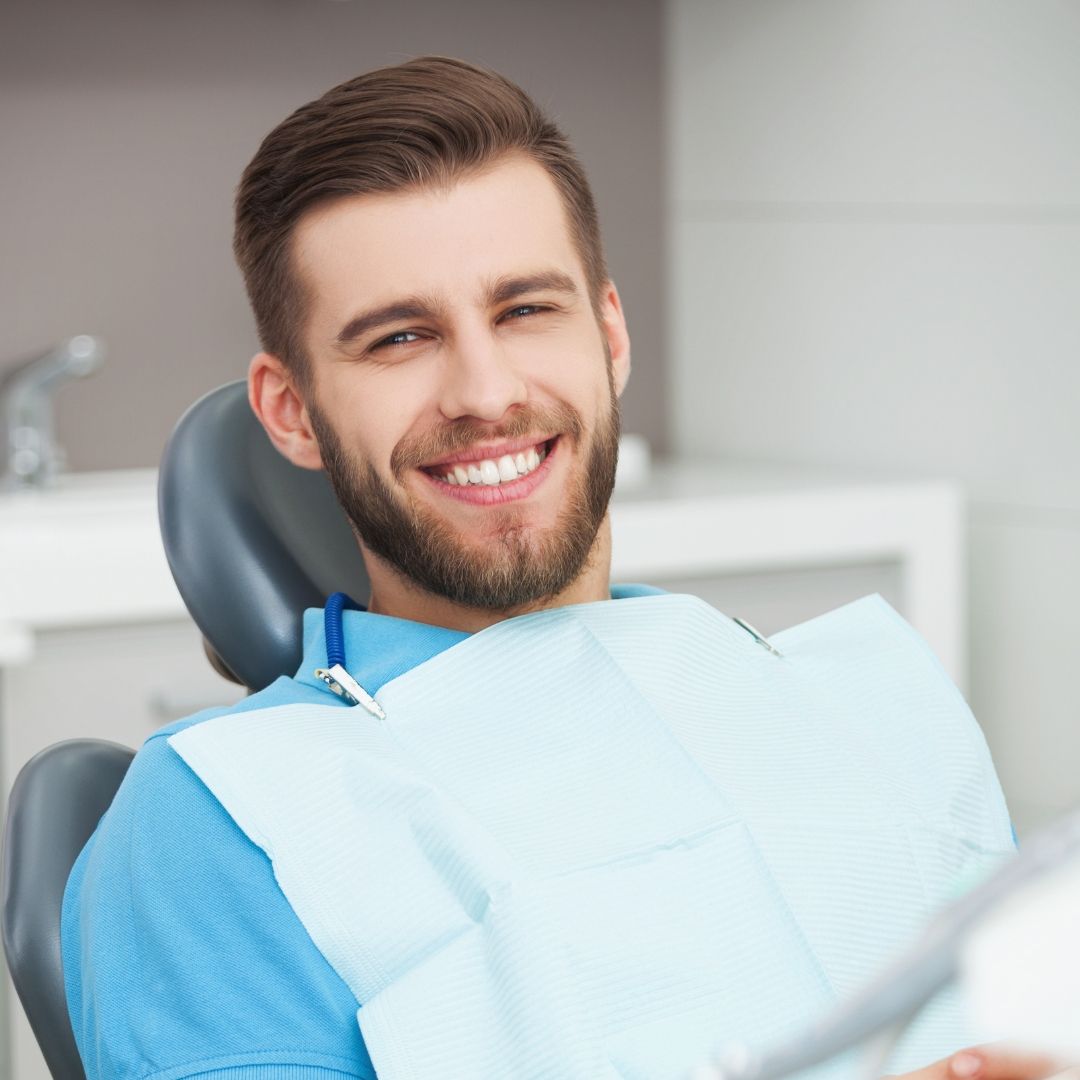 A smiling man with a beard in a dentist's chair, wearing a bib, with a bright, white background.