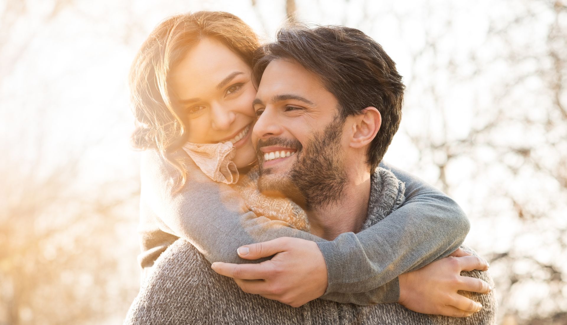 Woman hugging a smiling man from behind outdoors, sun shining. They appear happy.