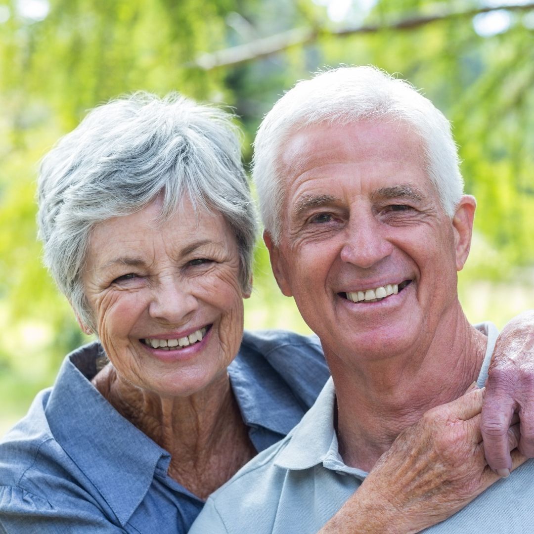 Smiling elderly couple embracing outdoors, bathed in natural light. Woman in blue shirt, man in grey.