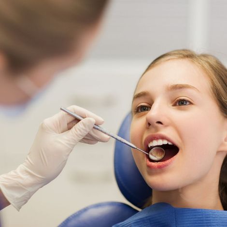 Dentist examining a smiling young woman's teeth with a mirror. The patient is seated in a dental chair.