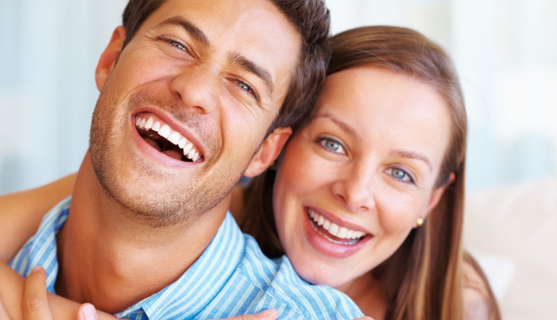 Smiling couple embracing, indoors. Man in blue shirt, woman with light eyes, both with bright smiles.