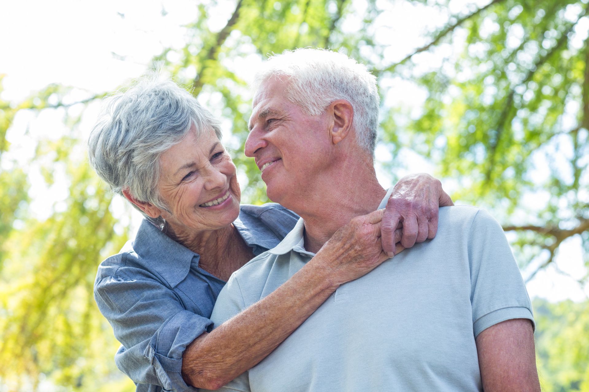Elderly couple embraces, smiling at each other outdoors. The woman has her arms around the man's shoulders. Sunlight shines through the trees.