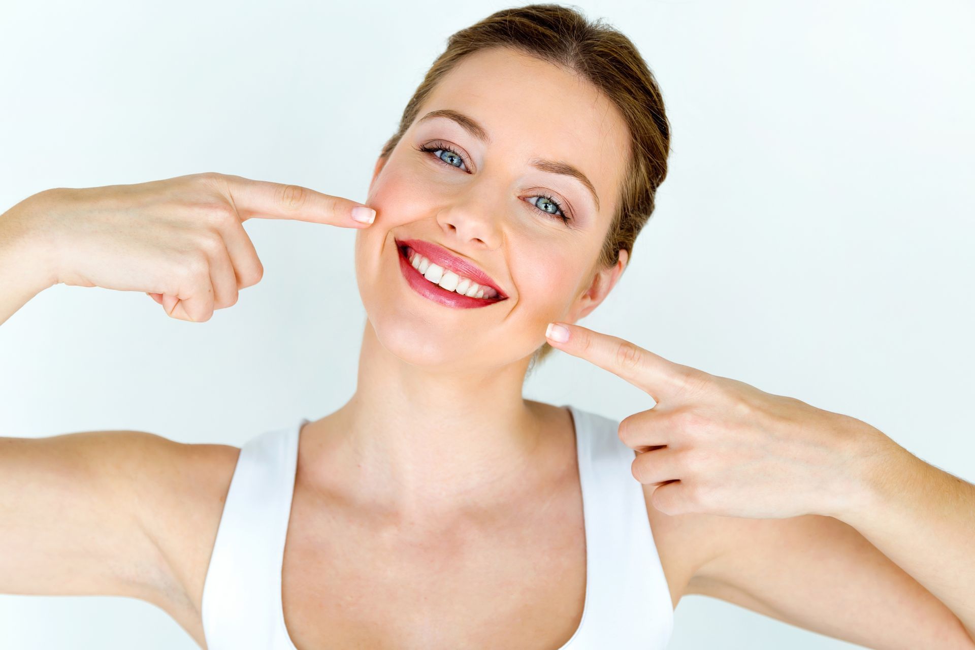 Woman with fair skin smiling, pointing to her teeth. She wears a white tank top against a white background.