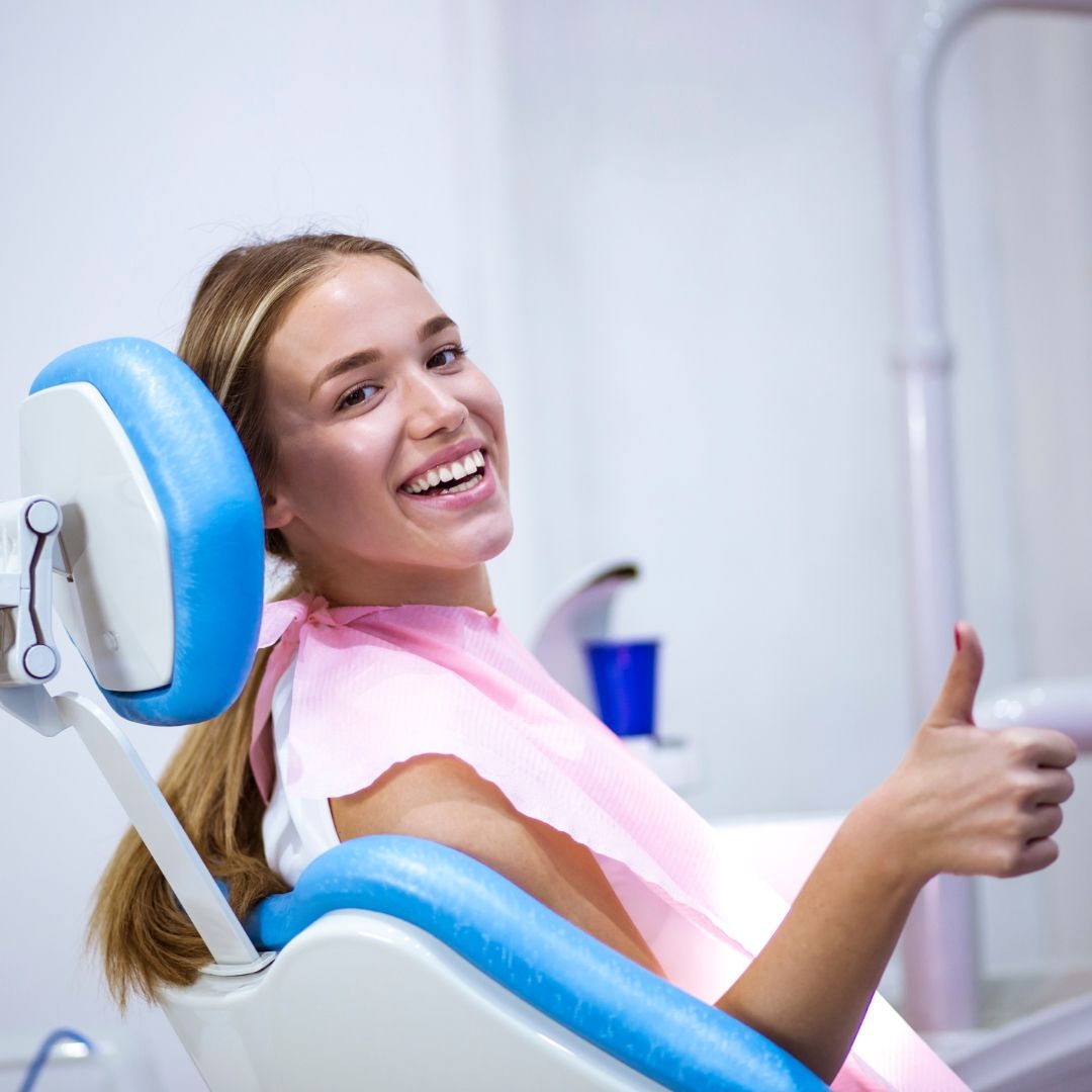 Woman in a dental chair smiling and giving a thumbs-up. She's wearing a pink bib and sitting in a blue and white chair.