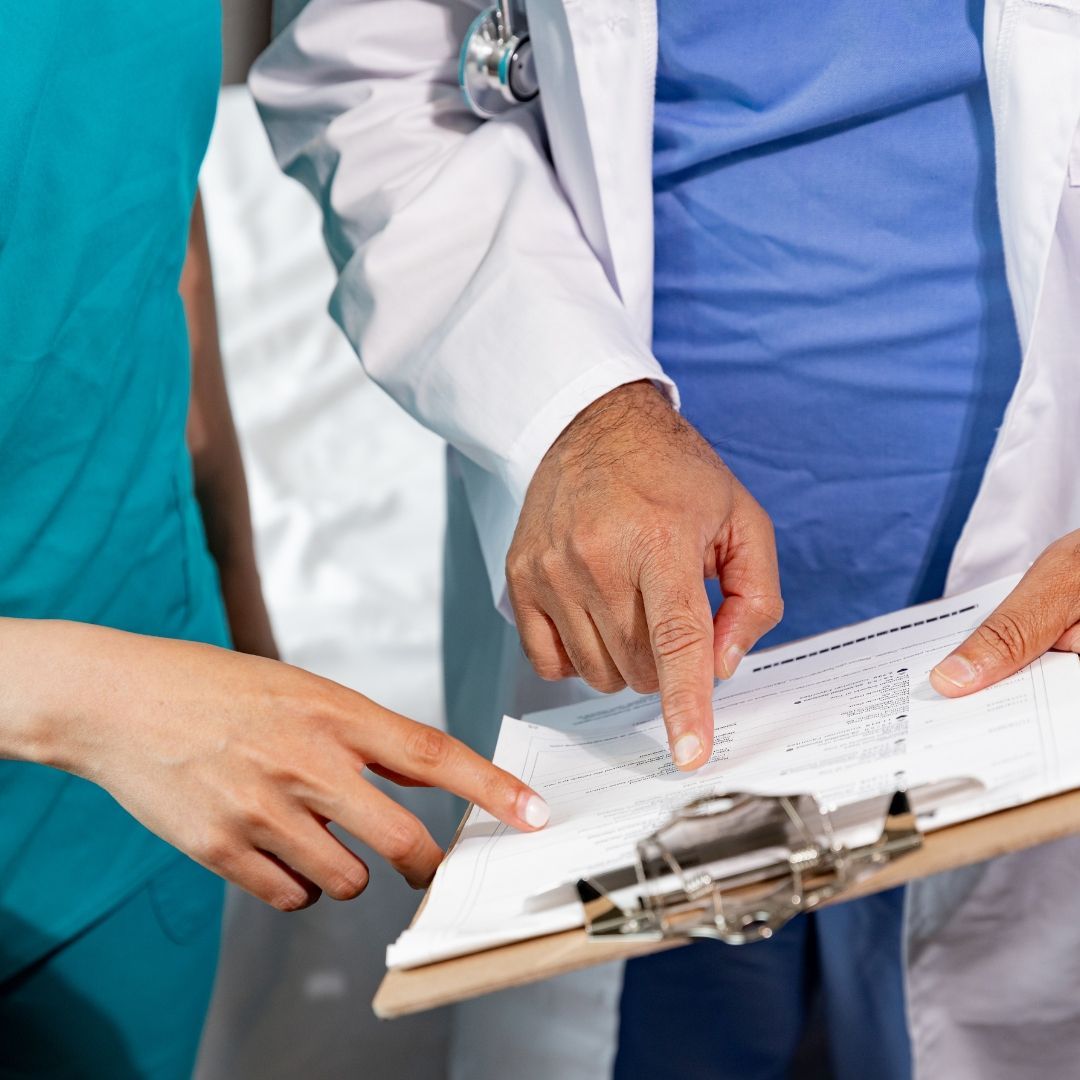 Doctor and nurse reviewing a medical chart on a clipboard, pointing and discussing data; setting appears to be a medical office.