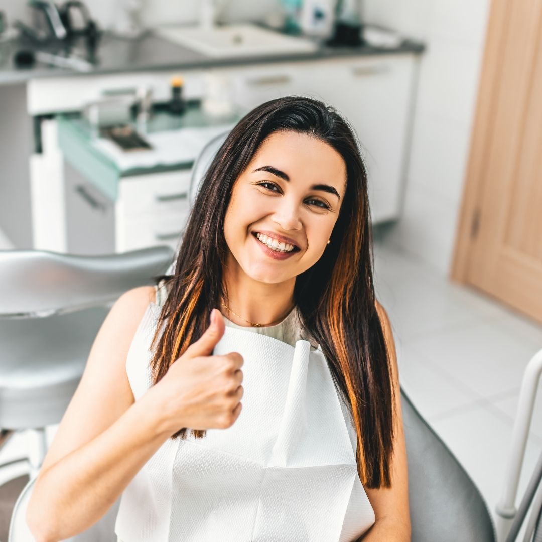 Woman in a dentist's chair with a white bib, smiling and giving a thumbs-up. She has long, dark hair in a dental office.