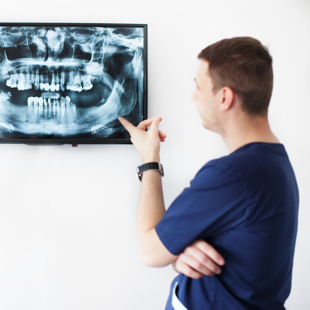 Dentist in blue scrubs pointing at a dental X-ray displayed on a white wall, explaining the results.