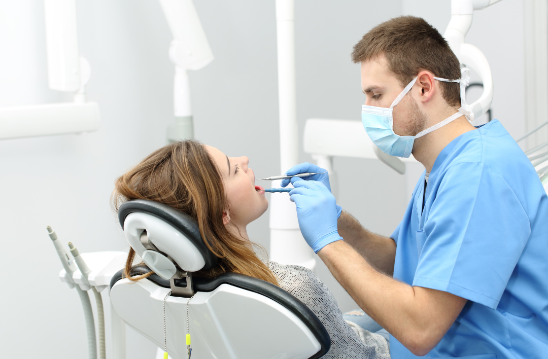 Dentist examining a patient's teeth in a dental office. The dentist wears a mask, gloves, and scrubs while using tools.