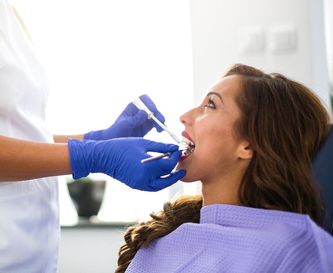 Woman in dentist chair, receiving a dental checkup. Dentist wearing blue gloves, uses tools in her mouth.