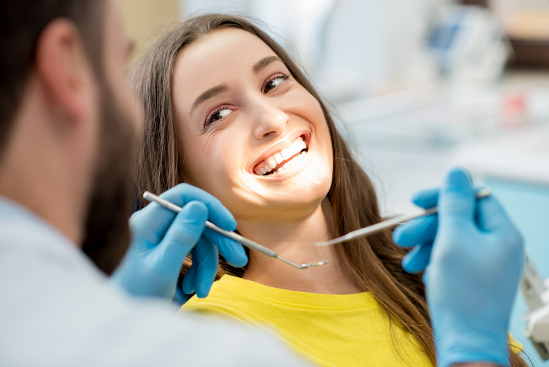 A dentist examining a smiling woman's teeth, using tools in a dental office. The woman wears a yellow shirt.