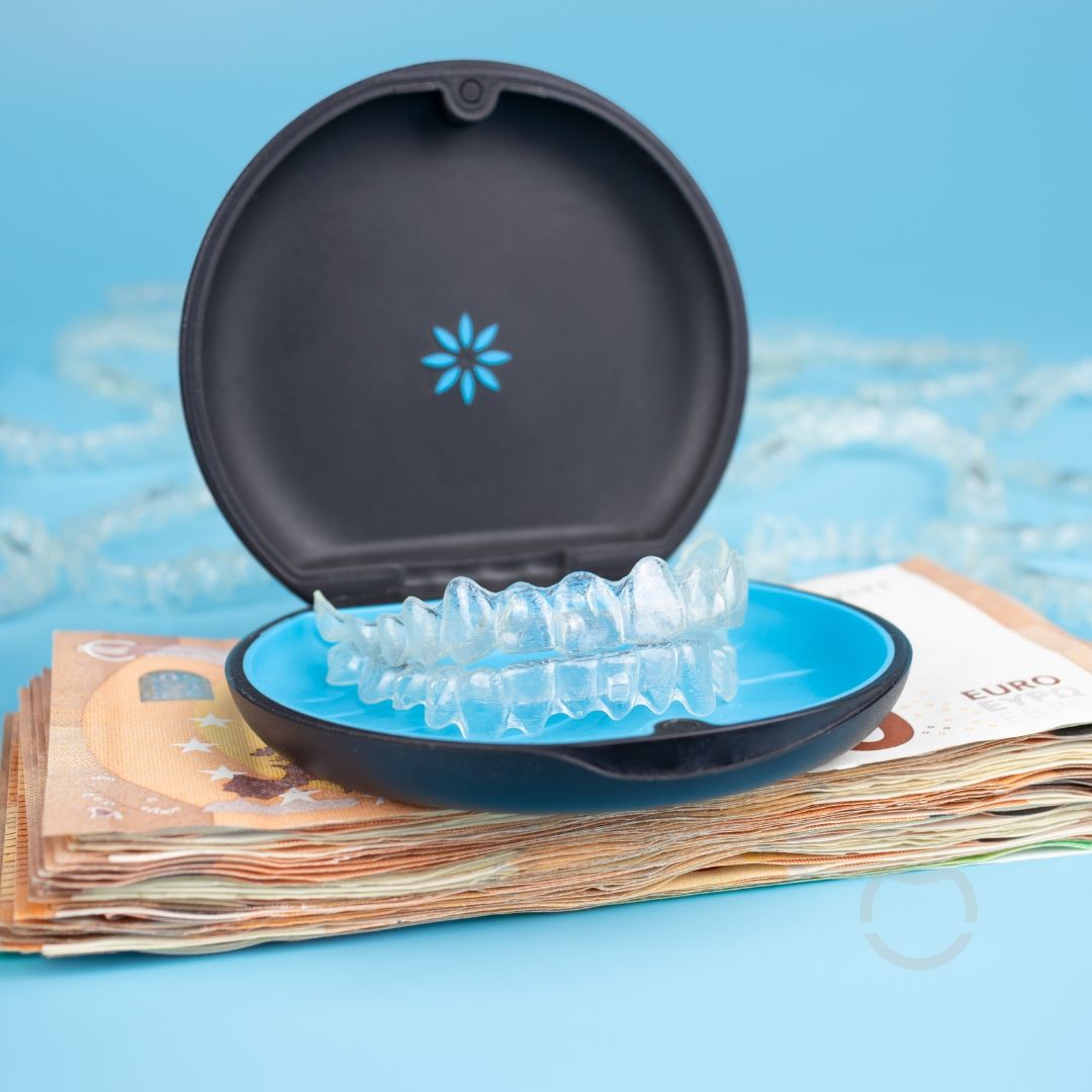 Clear aligners in a blue and black case sit on top of a stack of cash against a light blue background.