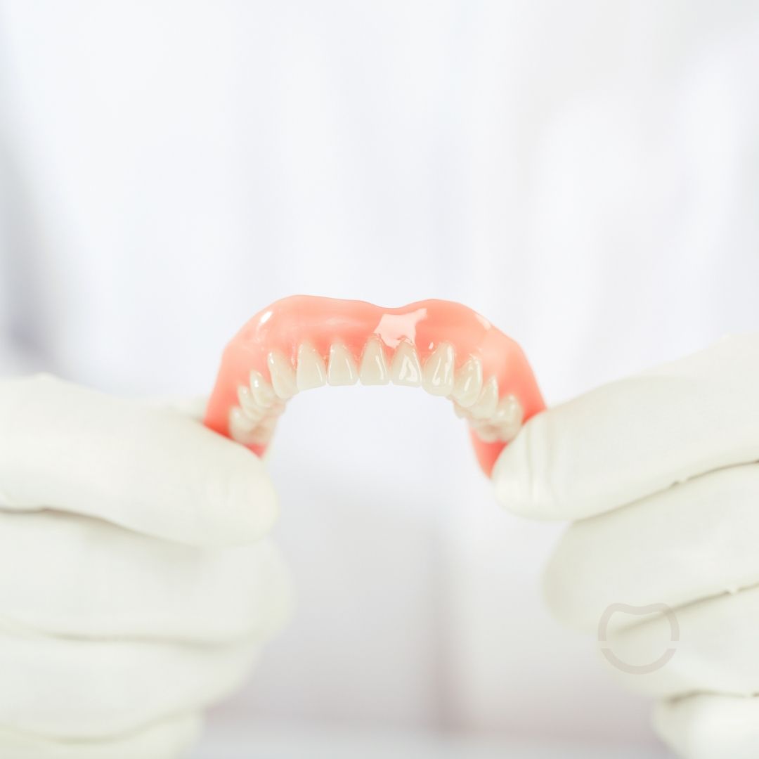 A person in white gloves holding a set of dentures against a white background. The dentures are pink with white teeth.