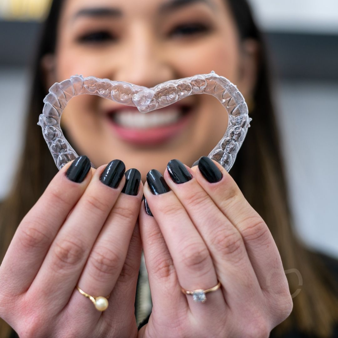 Woman holding clear aligners shaped like a heart, smiling with manicured nails, indoors.