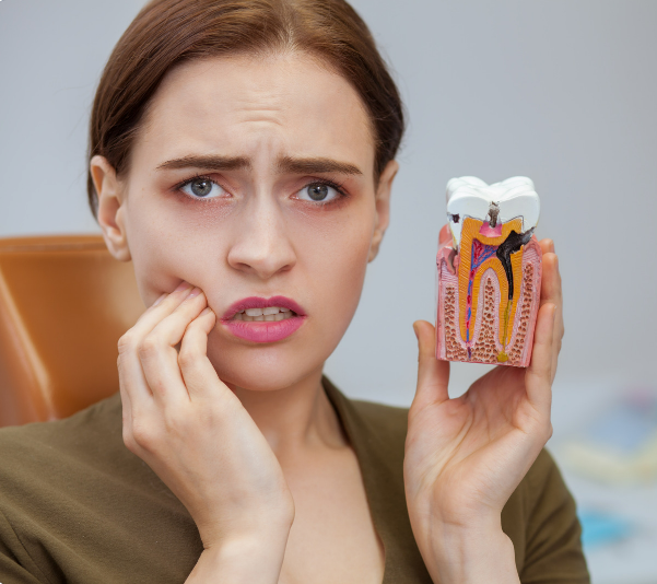 Woman in dental chair clutches cheek in pain, holding a tooth model showing a decayed tooth, grimacing.