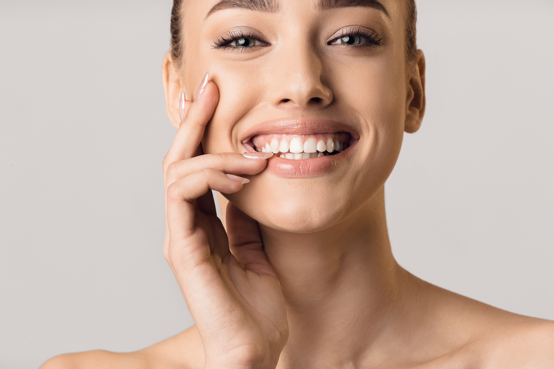 Woman with fair skin, smiling widely and touching her face, with a neutral background.