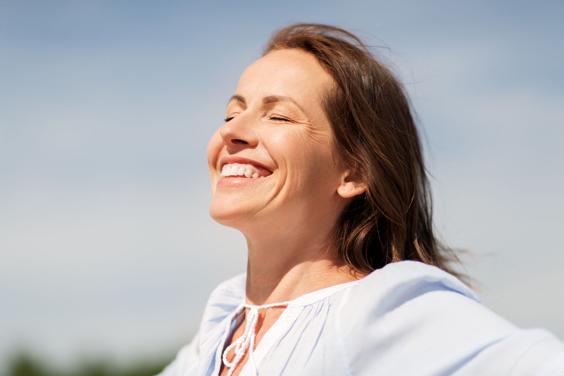Woman with brown hair smiles, eyes closed, outdoors with blue sky. Arms outstretched, wearing a light blue blouse.