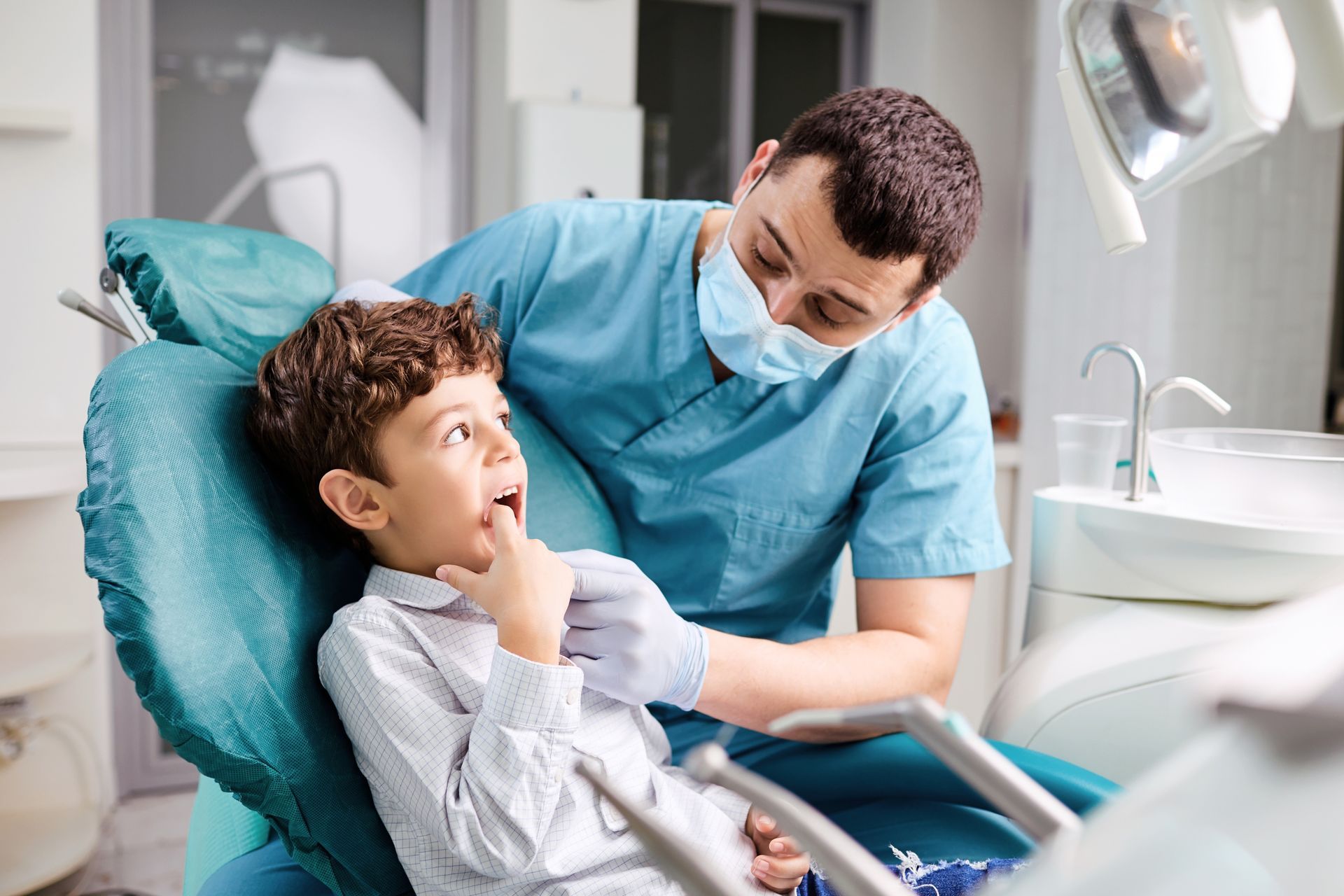 A dentist in blue scrubs and mask examines a young boy's teeth in a dental chair. The boy looks up with an open mouth.