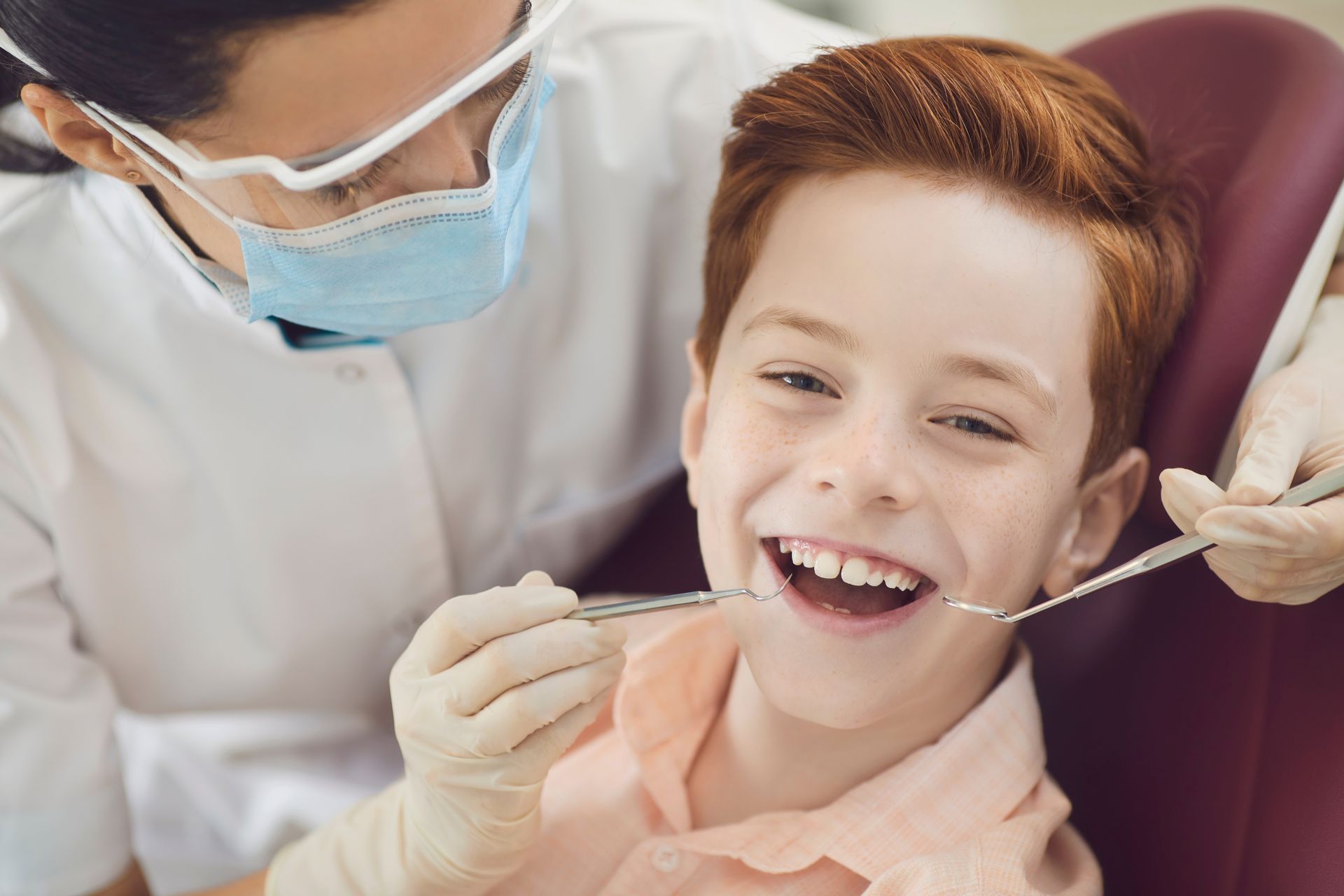 A dentist examines a smiling red-haired boy's teeth in a dental chair, using tools and wearing a mask.