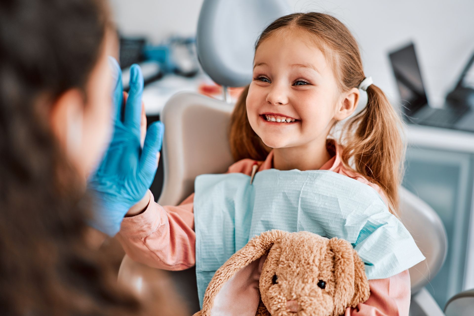 A young girl smiles brightly, giving a high five to a dentist in a clinic. She is holding a stuffed bunny.
