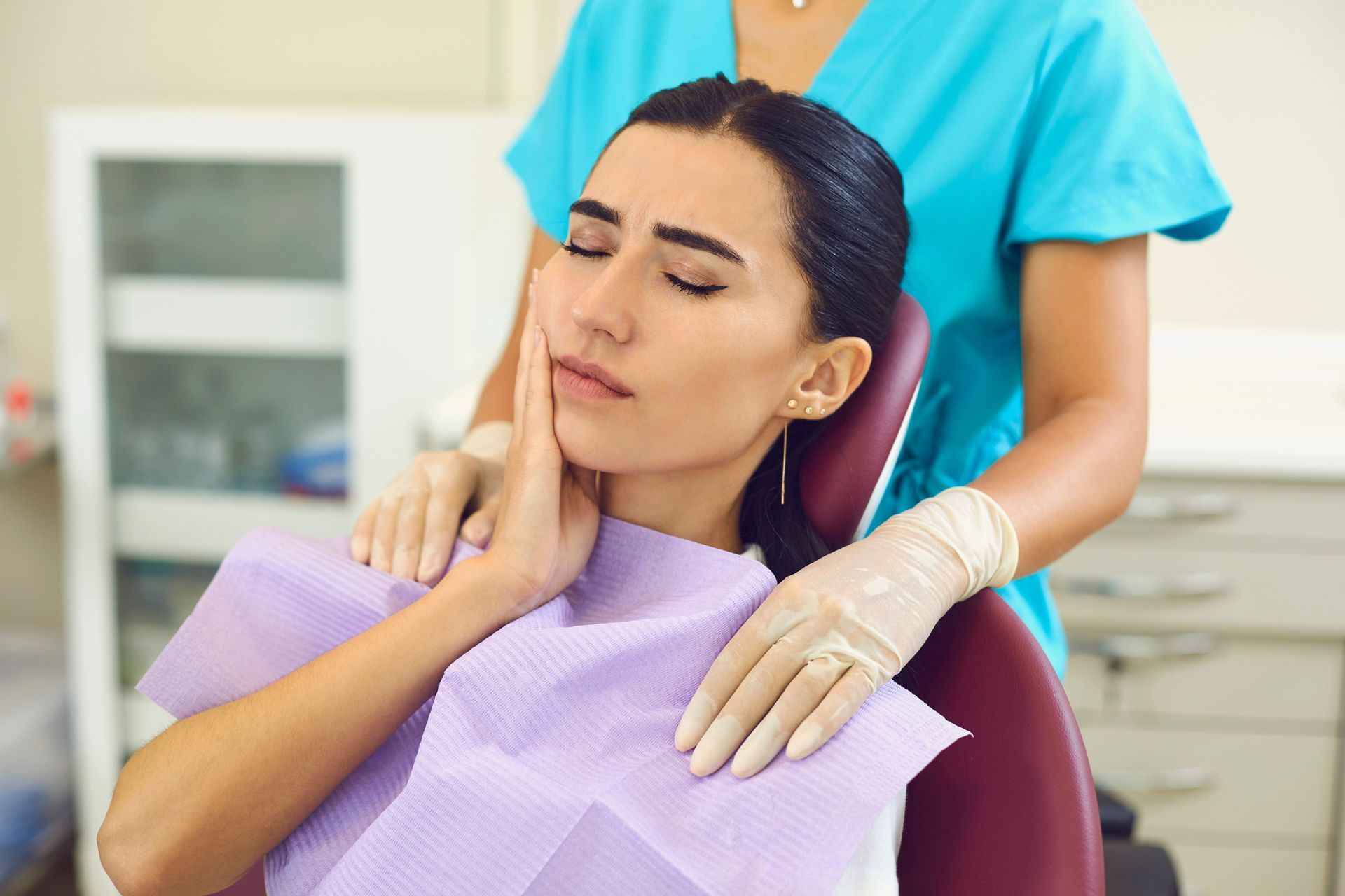 Woman in a dental chair holds her cheek, appearing in pain. A dental assistant in blue scrubs and gloves stands behind her.