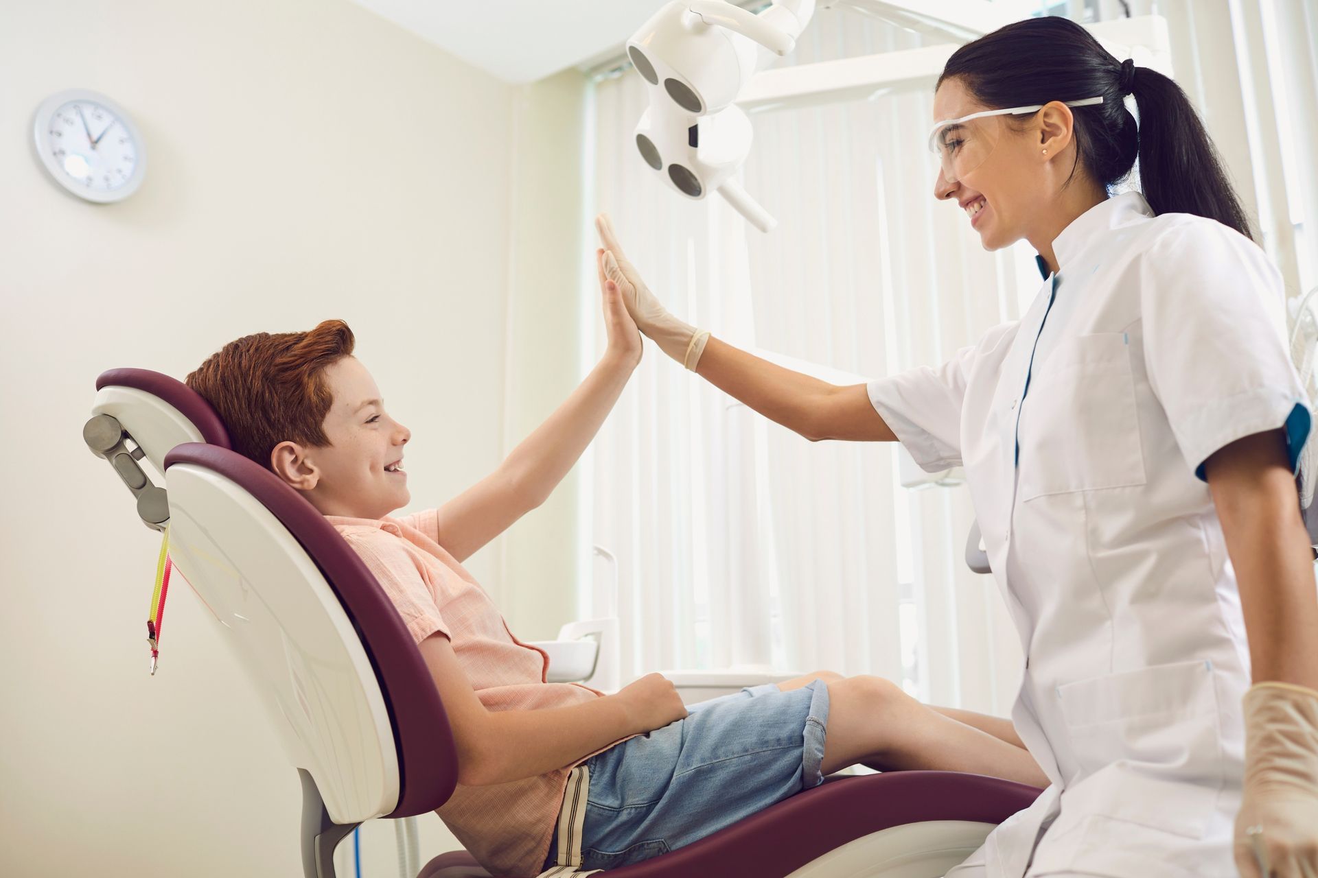 A dentist high-fives a smiling young boy in a dental chair. The boy has red hair and wears shorts; the dentist wears a lab coat.