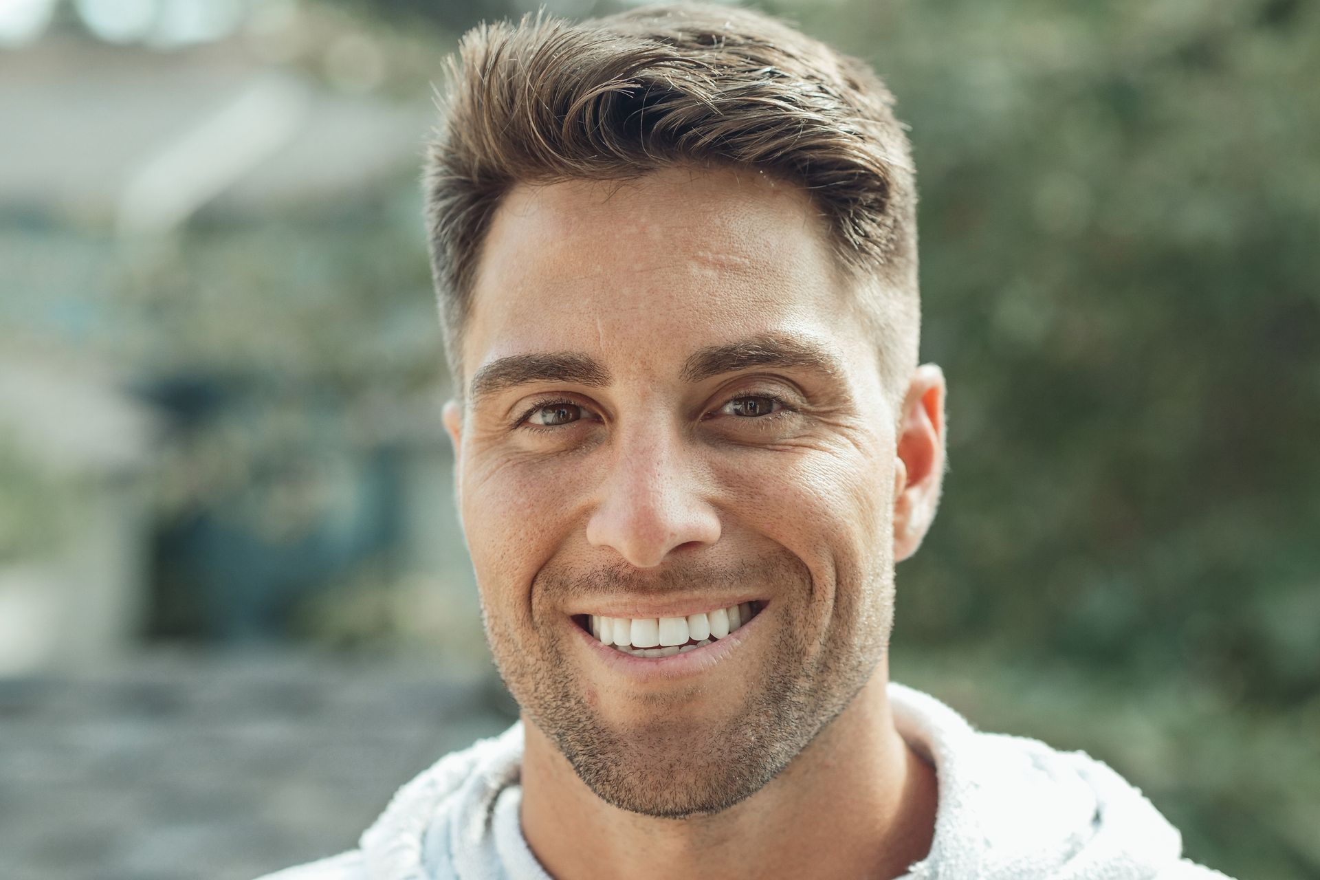 Smiling man with short brown hair, wearing a white hoodie, outdoors.